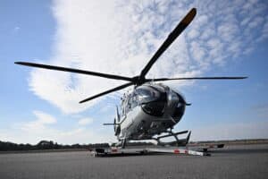 A gray helicopter is parked on a tarmac under a partly cloudy sky, with its rotor blades extended and positioned on a wheeled platform.