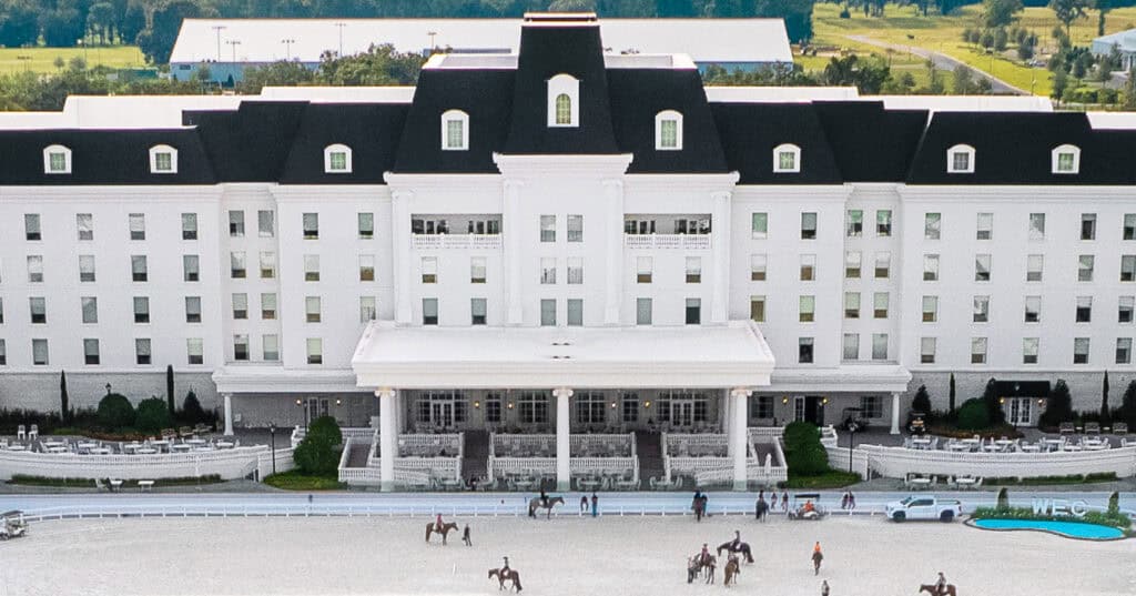 Large white hotel with black roof, symmetrical windows, and central entrance; people on horseback and walking are visible in the foreground near an outdoor arena.