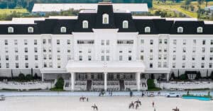 Large white hotel with black roof, symmetrical windows, and central entrance; people on horseback and walking are visible in the foreground near an outdoor arena.