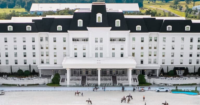 Large white hotel with black roof, symmetrical windows, and central entrance; people on horseback and walking are visible in the foreground near an outdoor arena.