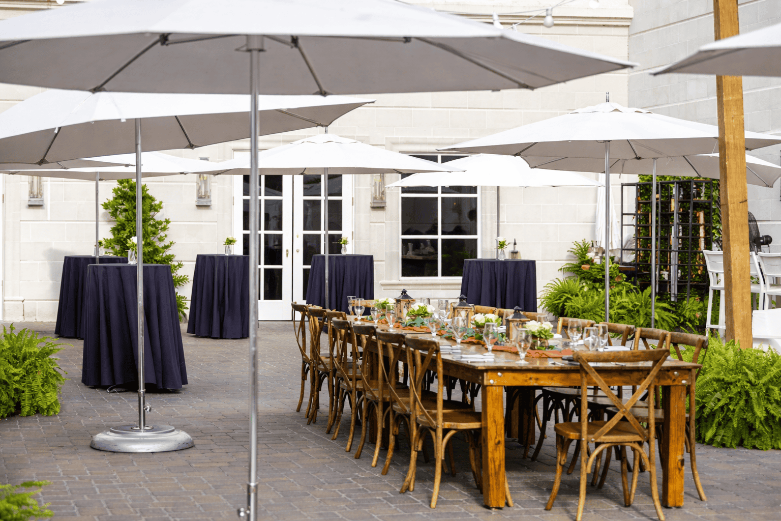 Outdoor patio with wooden dining table set for a meal, surrounded by wooden chairs and several tall round cocktail tables under large white umbrellas.