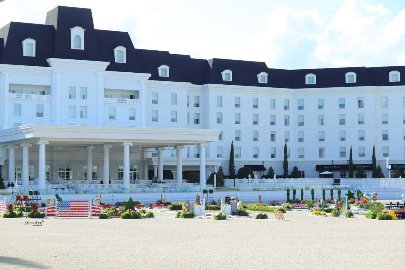 A large white building with a dark roof stands behind an outdoor equestrian arena set up with various colorful jumps and decorative greenery.