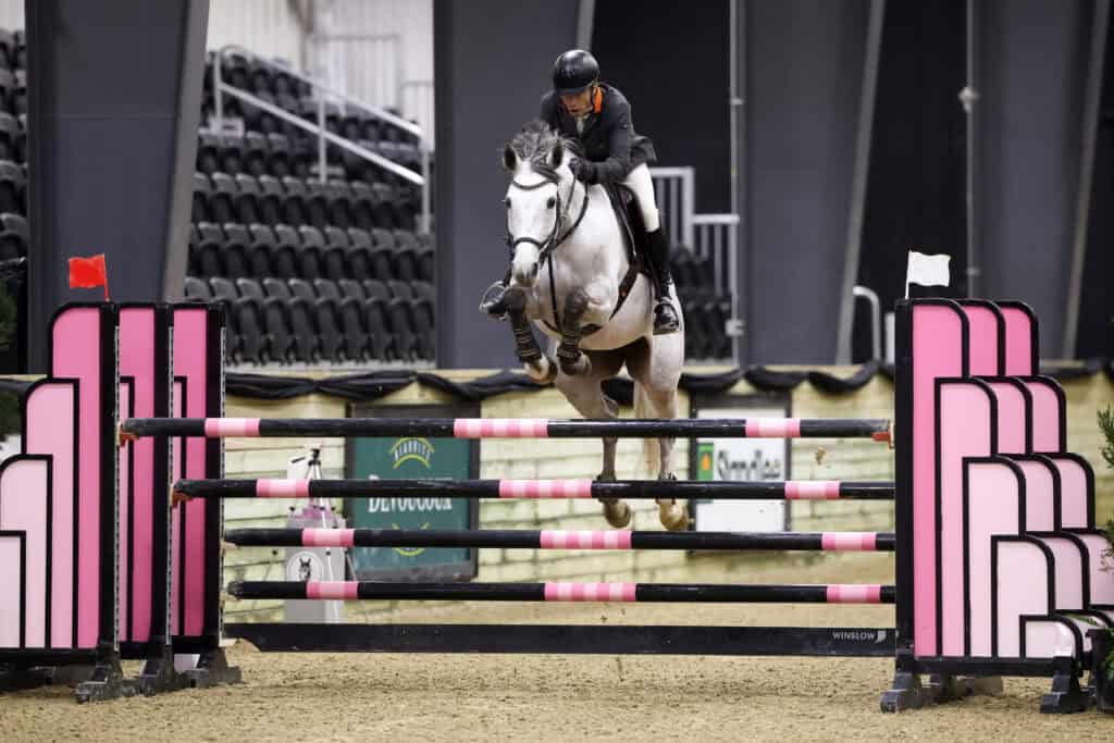 A rider wearing a black helmet and jacket guides a gray horse over a pink and black jump in an indoor equestrian arena.