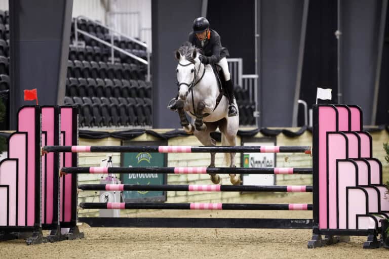 A rider and horse clear a jump at an outdoor equestrian event, with a large white building in the background and "First Equestrian" signage visible.