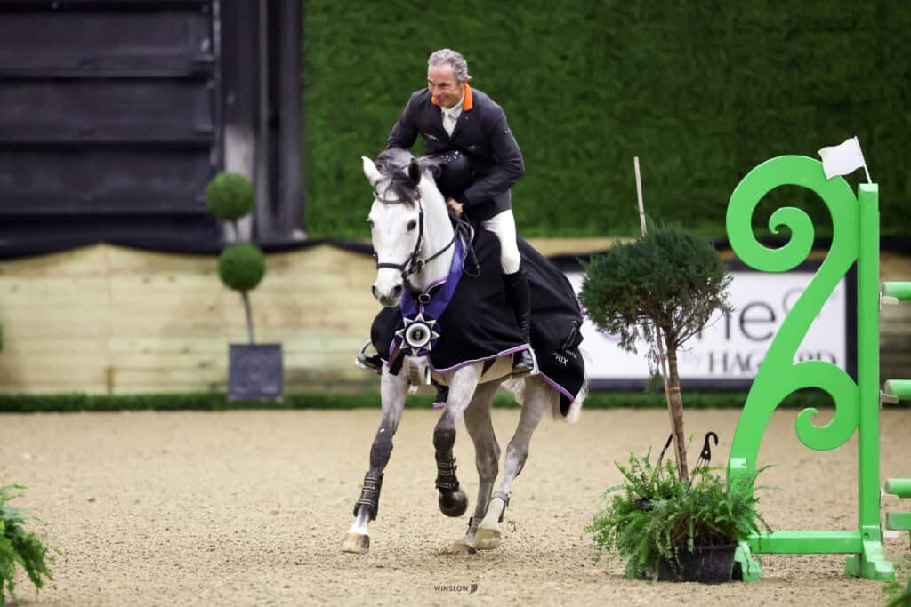 A man in formal riding attire rides a grey horse covered with a black blanket inside an indoor arena, passing green decorative jumps and plants.