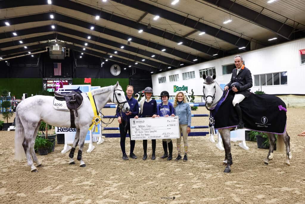 Five people stand indoors holding a large check between two white horses wearing ribbon rosettes and blankets, with jumps and equestrian event signage in the background.