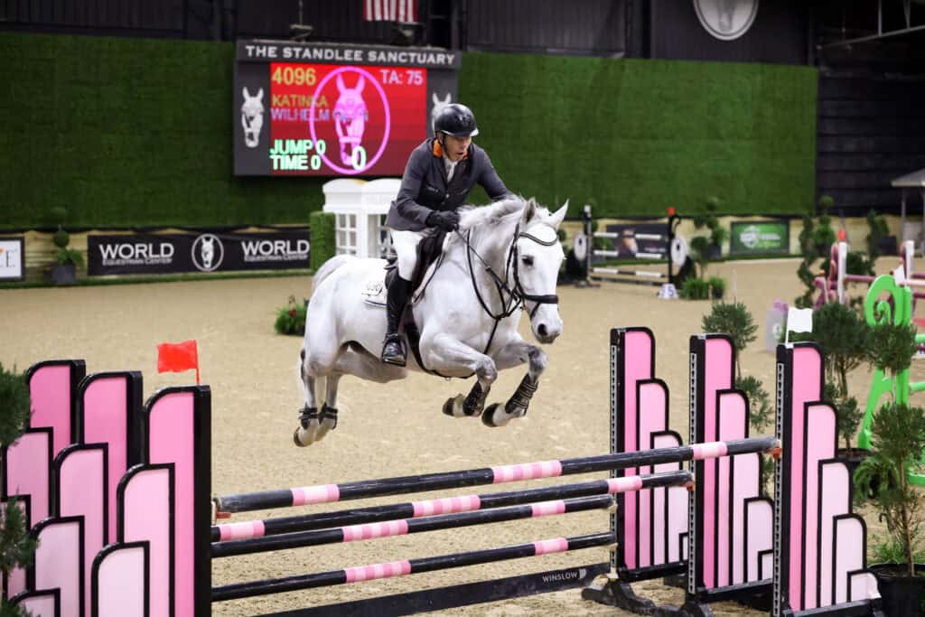 A rider on a gray horse jumps over a pink and black obstacle during an indoor equestrian event, with a scoreboard visible in the background.