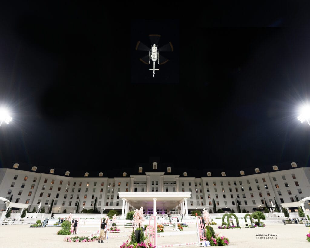 A large white building is illuminated at night with people and equestrian jumps set up in the foreground; a drone hovers in the sky above.