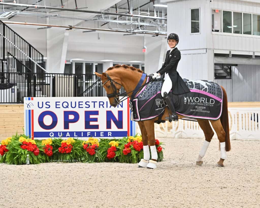 A rider in formal attire sits on a decorated horse in an indoor arena beside a sign reading "US Equestrian Open Qualifier." The horse wears a winner's sash and blanket.