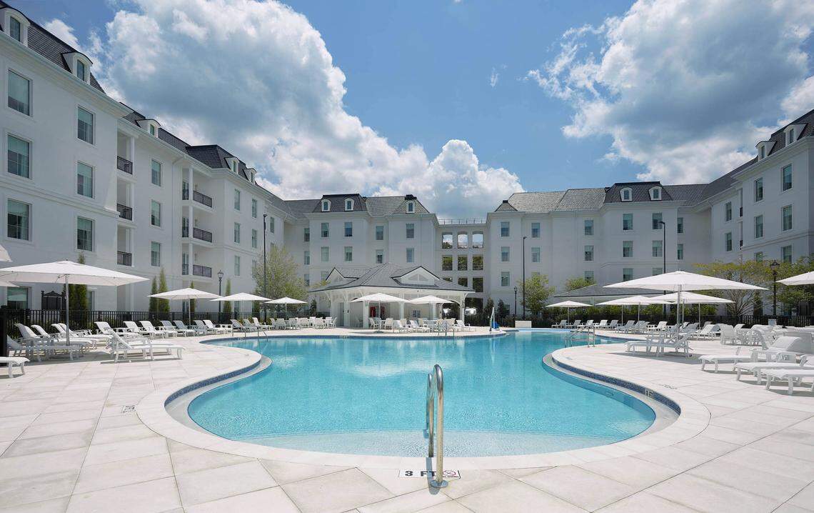Large outdoor pool with white poolside lounge chairs and umbrellas, surrounded by a multi-story white hotel building under a partly cloudy sky.