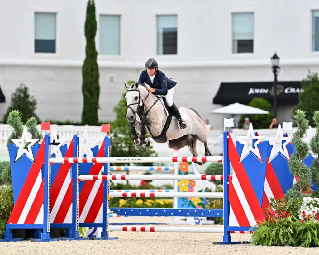 A rider in equestrian gear jumps a gray horse over a red, white, and blue star-decorated obstacle in a show jumping competition.