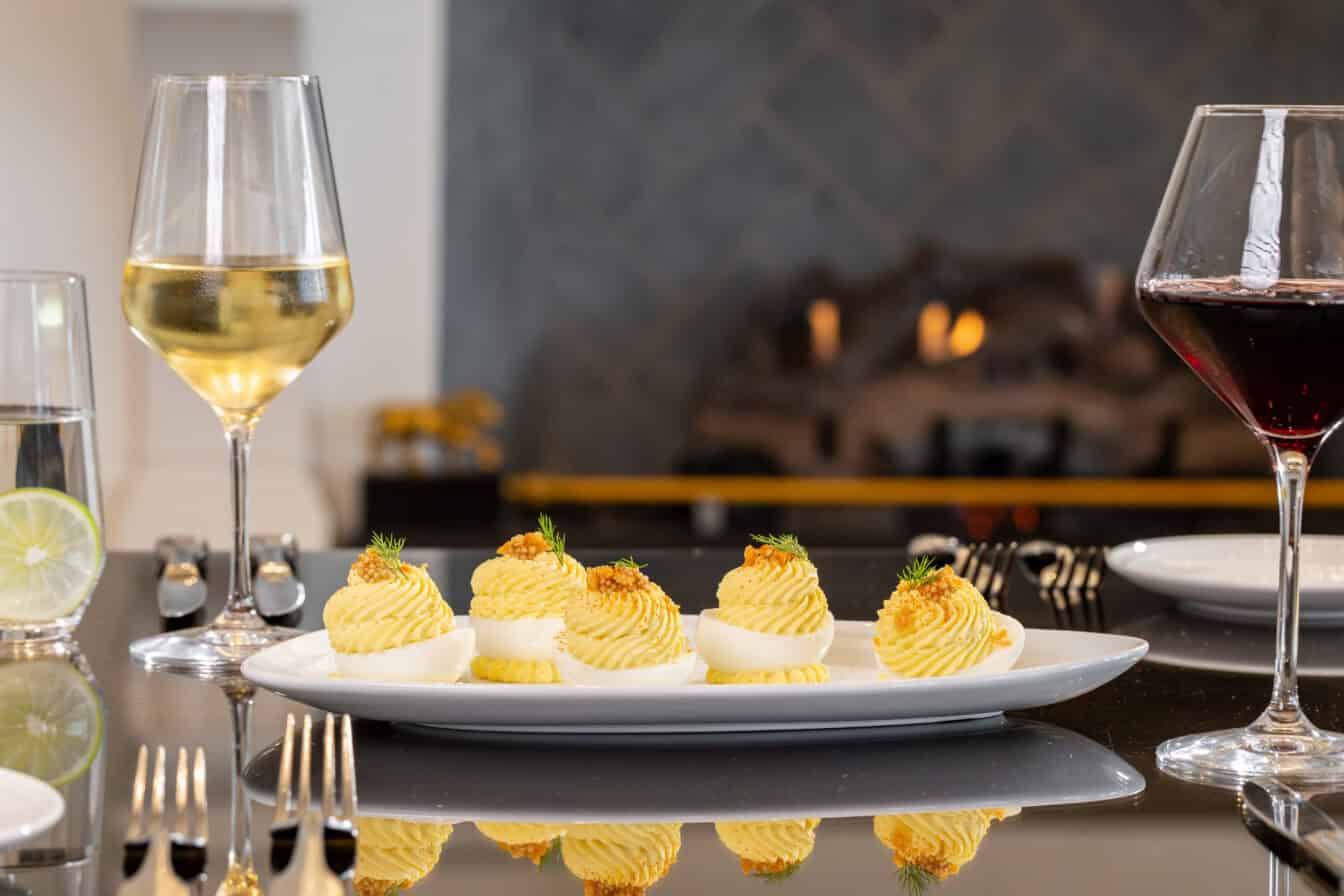 A plate of deviled eggs garnished with herbs is set on a table with wine glasses, silverware, and a fireplace in the background.