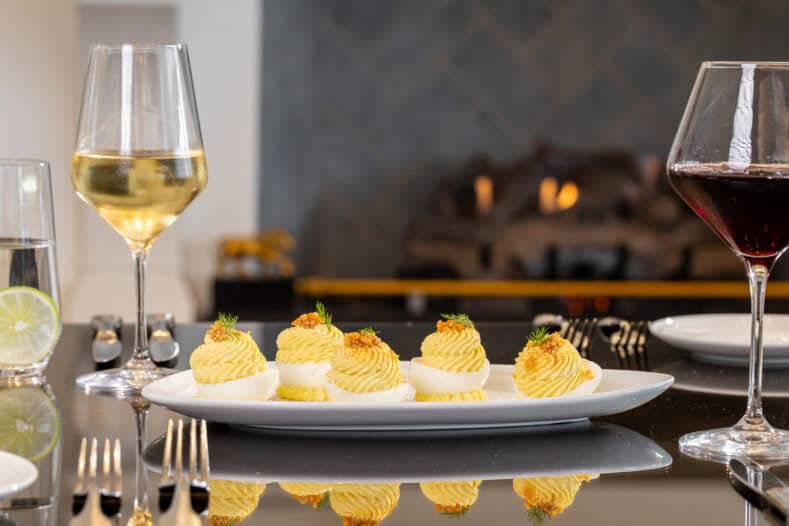 A plate of deviled eggs garnished with herbs is set on a table with wine glasses, silverware, and a fireplace in the background.