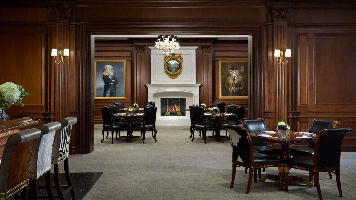 Elegant dining area with dark wood paneling, black leather chairs, tables set for service, a lit fireplace, chandelier, and two large portraits on either side of a mantel mirror.