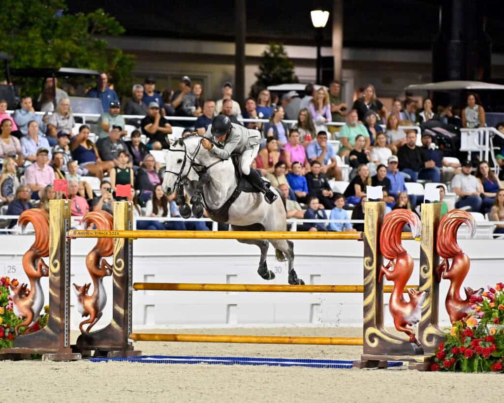 A rider on a gray horse jumps over a decorative obstacle in an equestrian event, with a crowd of spectators watching in the background.