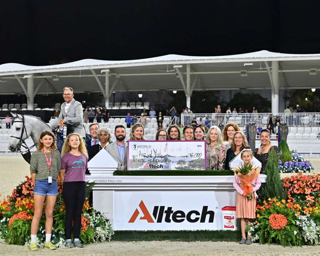 A group of people, a horse, and a large check stand together in front of an "Alltech" sign at an outdoor equestrian event, with flowers and stadium seating in the background.