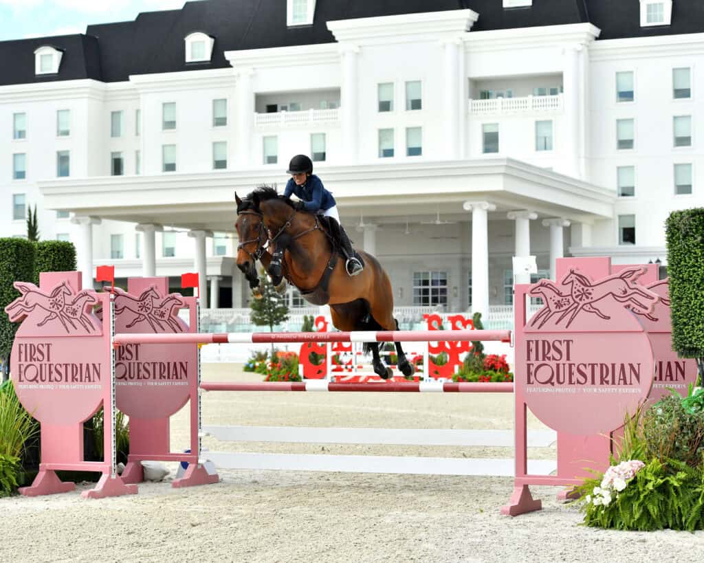 A rider and horse clear a jump at an outdoor equestrian event, with a large white building in the background and 