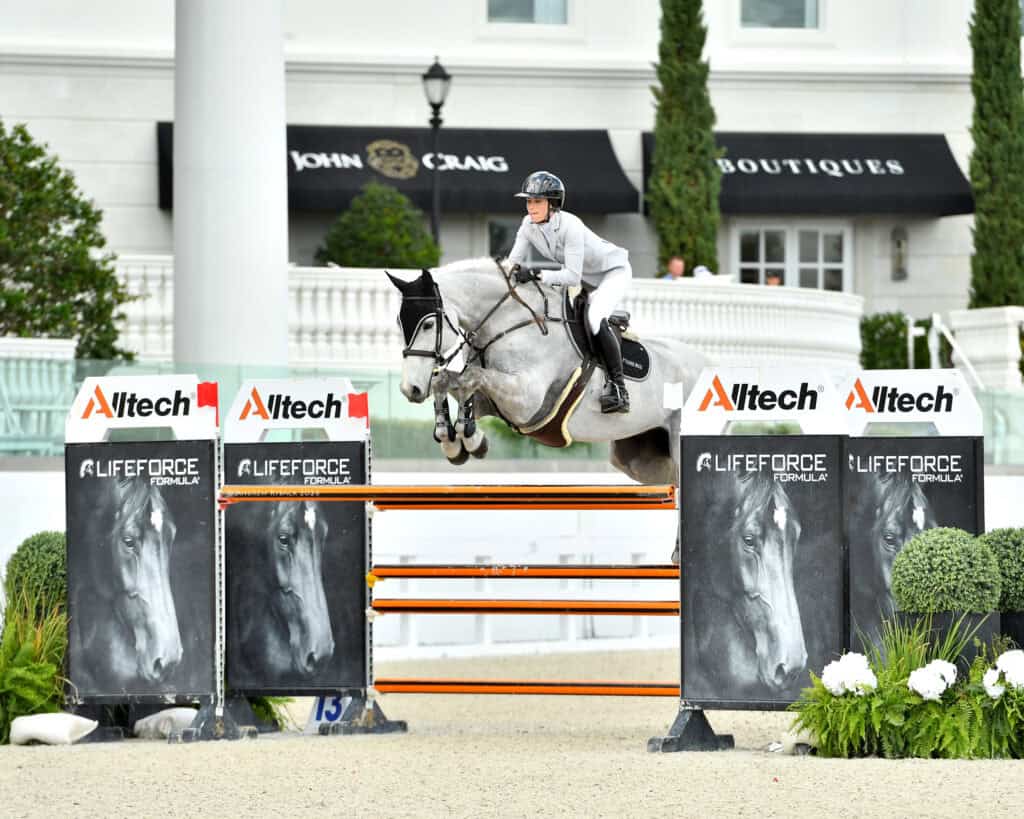 A rider in a white jacket and helmet guides a gray horse over a jump during an equestrian competition, with branding and greenery visible in the background.