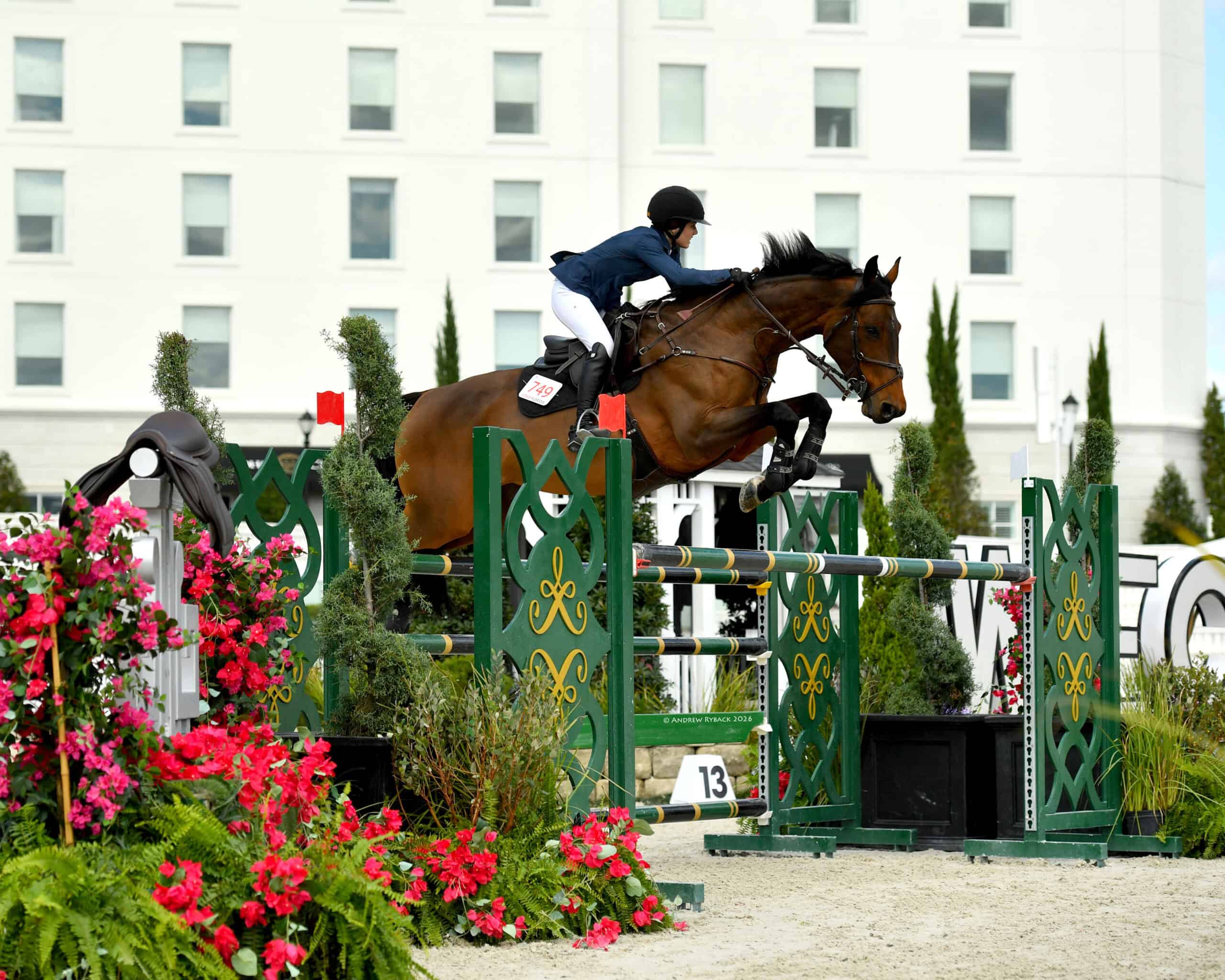 A rider on a brown horse jumps over a green and yellow obstacle during an equestrian event, with a building and flowers in the background.