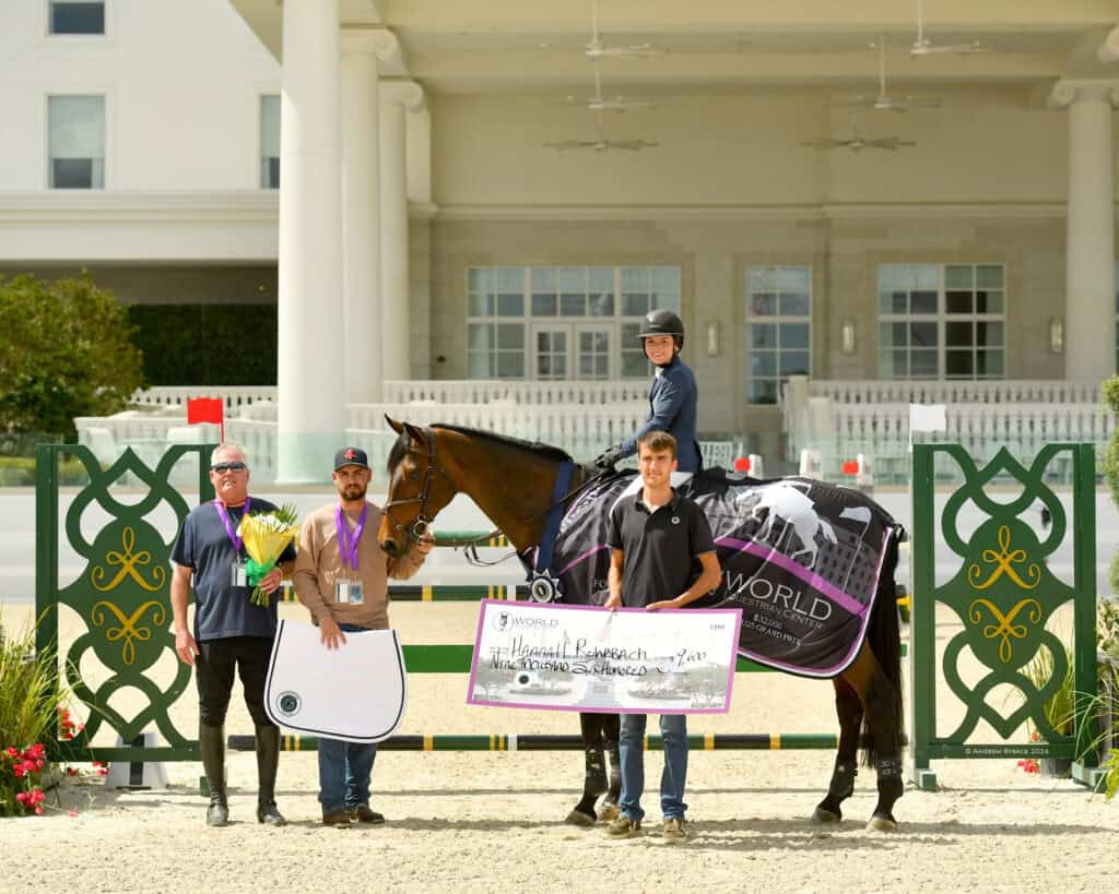 A rider on a horse holds a large check, accompanied by three people, flowers, and equestrian gear, in front of a building and decorative green gates.