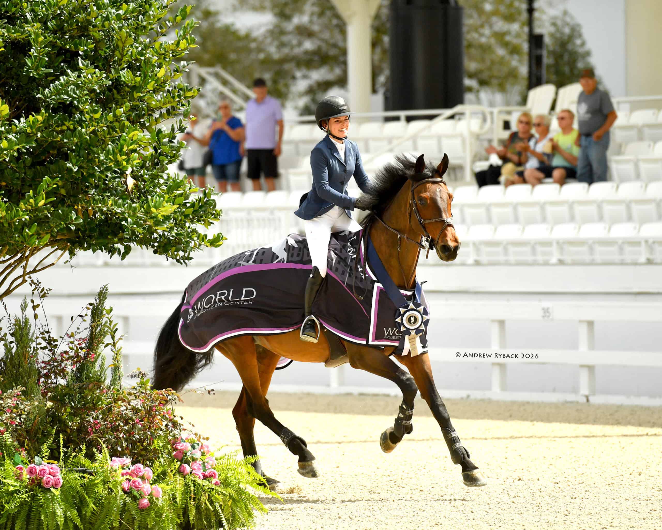 A rider in a helmet and blue jacket guides a horse draped in a winner's blanket and medal through a show arena; spectators watch from white bleachers.