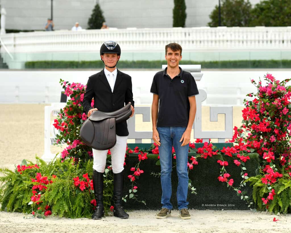 Two men stand side by side in front of a flower display; one wears equestrian attire and holds a saddle, while the other is casually dressed in a polo shirt and jeans.