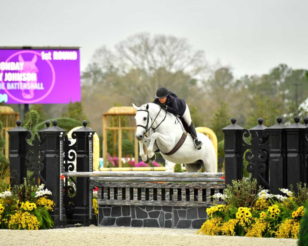A rider in equestrian attire jumps a white horse over a fence during a show jumping competition, with flowers and a scoreboard in the background.