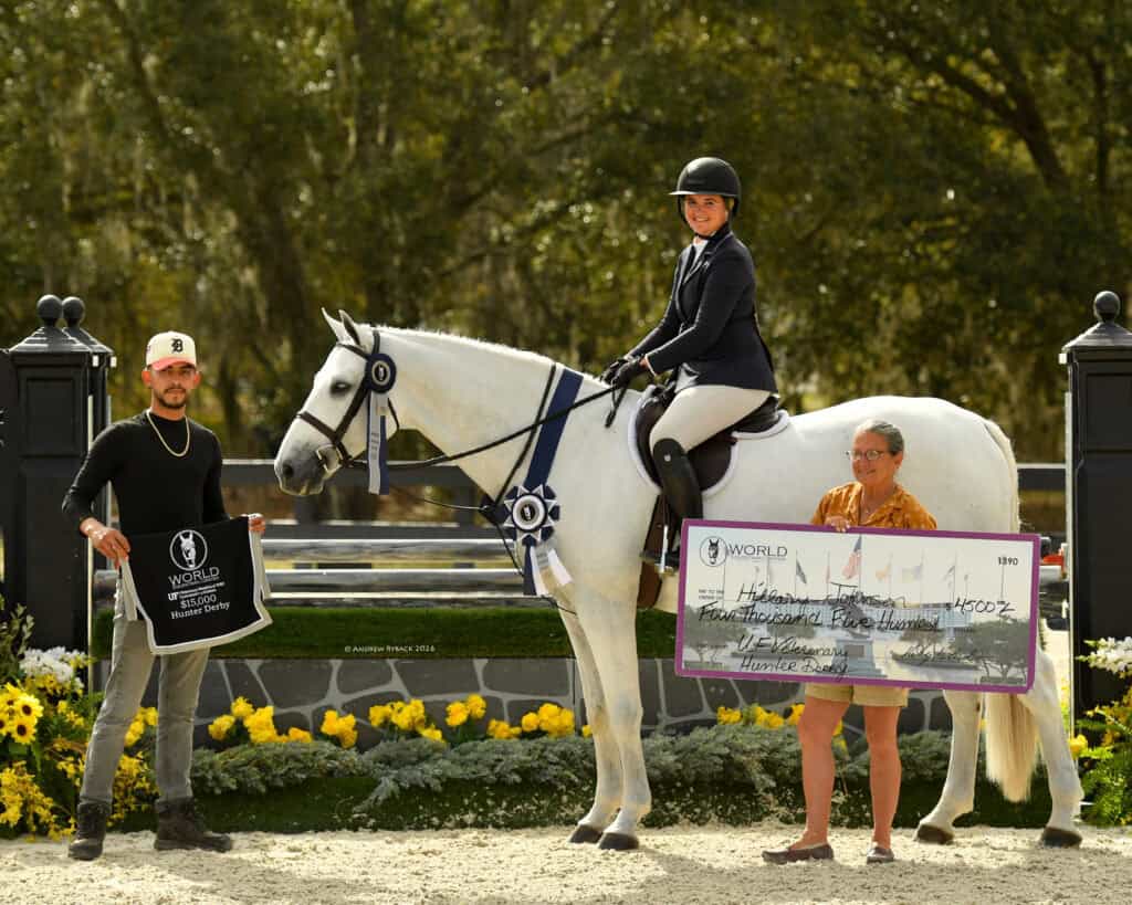 A rider on a white horse poses with two people holding a banner and an oversized check in an outdoor equestrian arena.