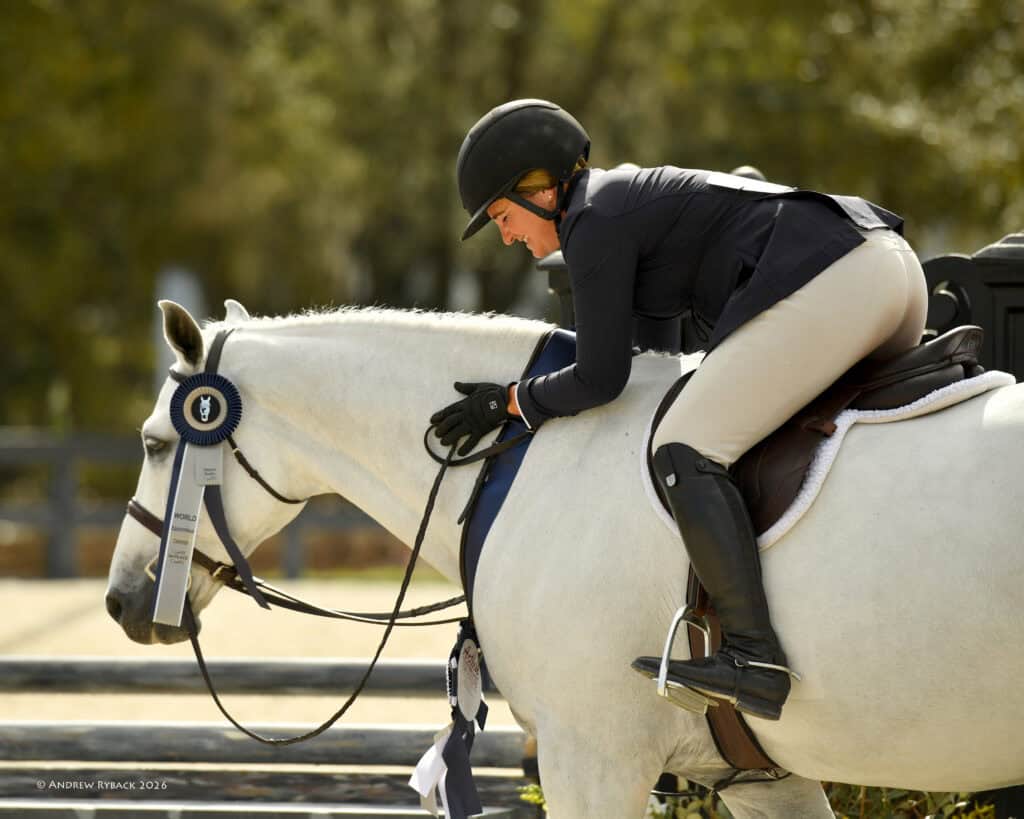 Equestrian rider in show attire leans forward to embrace a white horse wearing two award ribbons, outdoors in a riding arena.