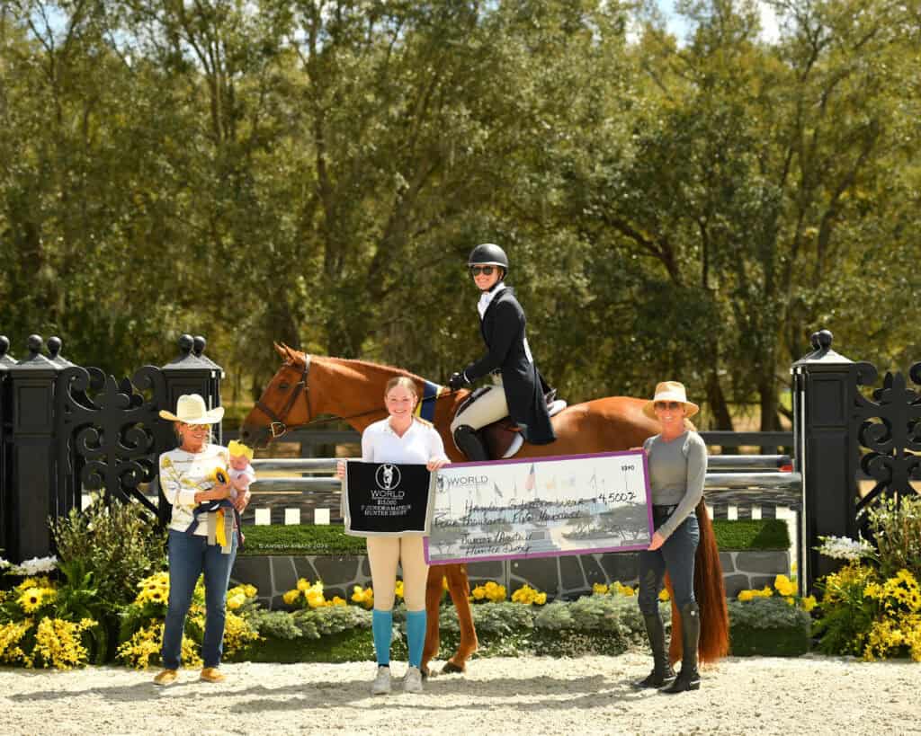 Four people and a horse at an outdoor equestrian event; one person on horseback and three others holding a large check in front of a decorative gate and flowers.