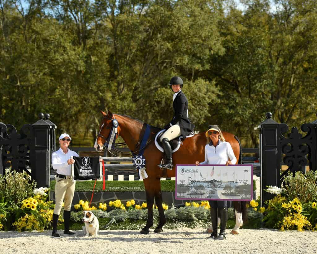 Three people and a dog pose outdoors with a horse; one person on horseback, the others standing with a large check and ribbon in front of a decorated equestrian event backdrop.