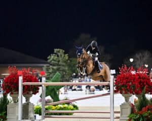 Equestrian rider and horse jump over a red and white obstacle during a nighttime show jumping competition, surrounded by flower arrangements.