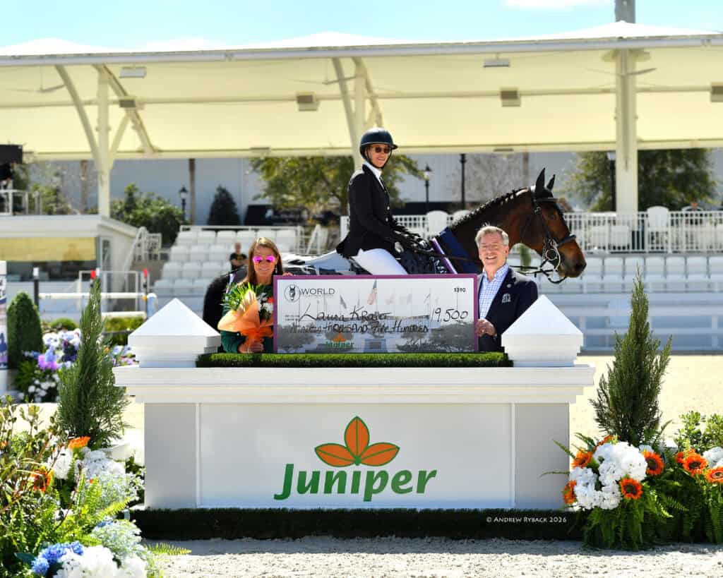 A rider on horseback and two people stand behind a large ceremonial check at an outdoor equestrian event, with flowers and a Juniper sign in front.