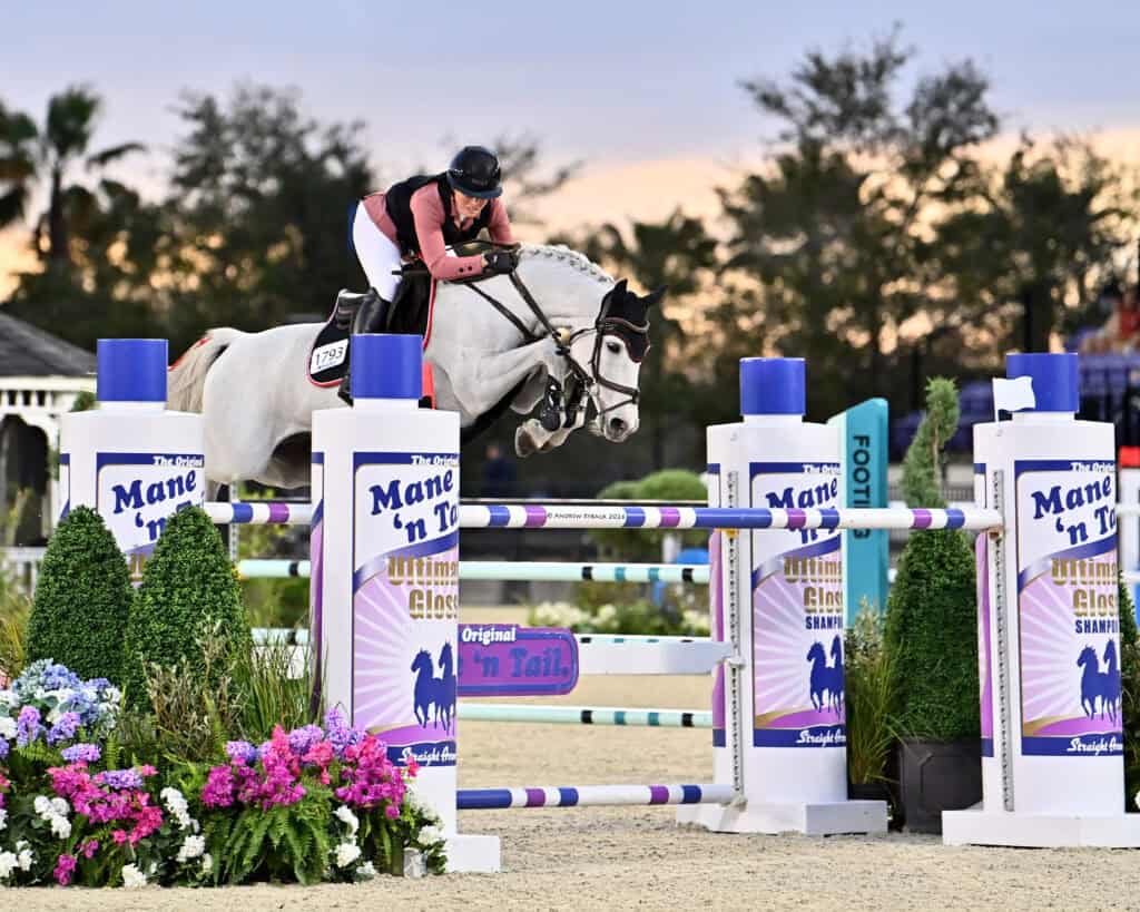 A rider on a white horse jumps over an obstacle during an equestrian event, with flowers and sponsor banners visible in the arena.