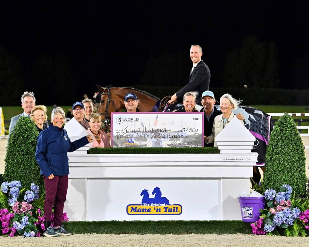 A group of people pose with a horse and rider holding a large award check for $37,500 at an outdoor equestrian event, standing behind a decorated jump.