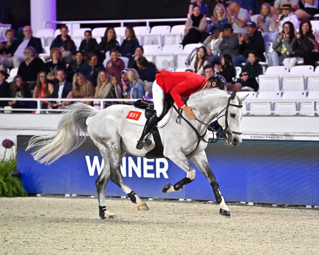 A rider in a red jacket leans forward to hug a gray horse during an equestrian event, with spectators seated in the background and a "WINNER" sign visible.