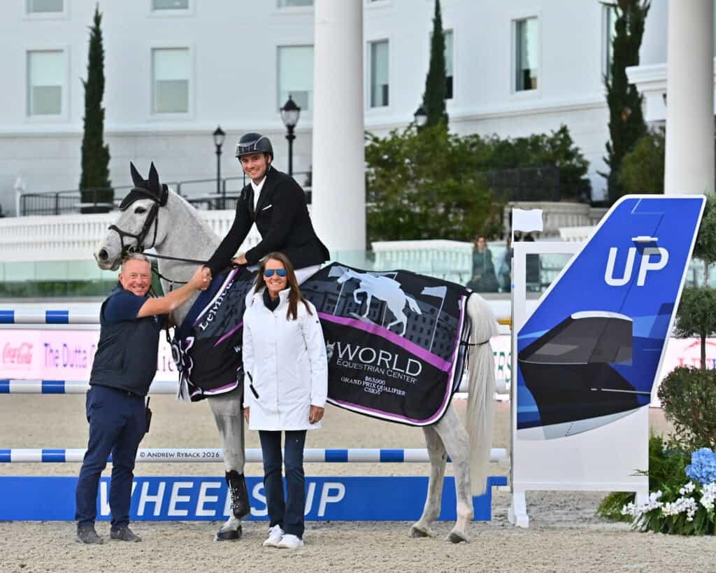 A rider on a white horse poses with two people beside a jump bar and a large "UP" sign at an equestrian event.