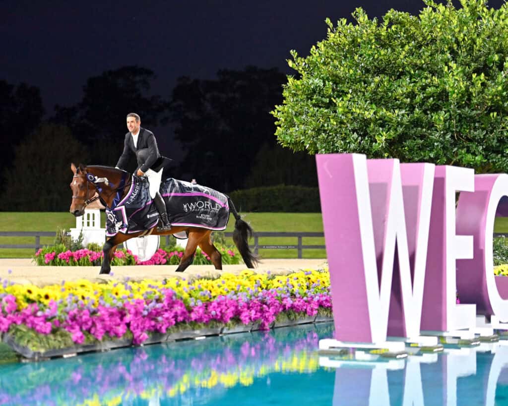 A rider on a horse draped in a winner’s blanket passes a large "WEC" sign next to a reflective pool, with flowers and greenery in the background at night.