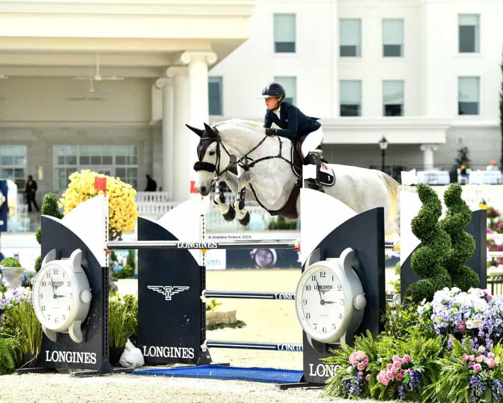Equestrian rider in helmet and uniform jumps a grey horse over a Longines-branded obstacle during an outdoor show jumping competition.