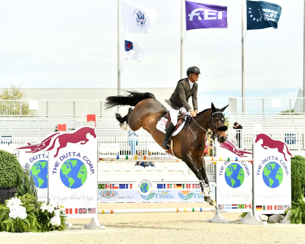 A rider on horseback jumps over an obstacle during an equestrian competition, with flags and empty stands visible in the background.