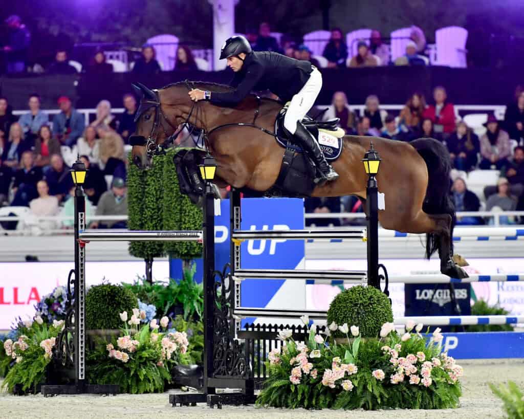 A rider and horse in mid-jump over an obstacle during an equestrian event, with spectators watching from the stands.