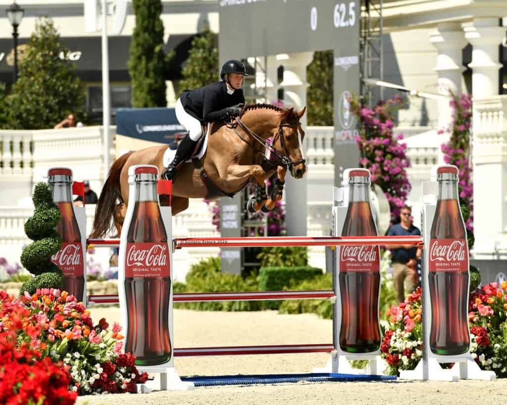 A person in equestrian gear rides a horse over a jump decorated with large Coca-Cola bottle images at an outdoor show jumping event.