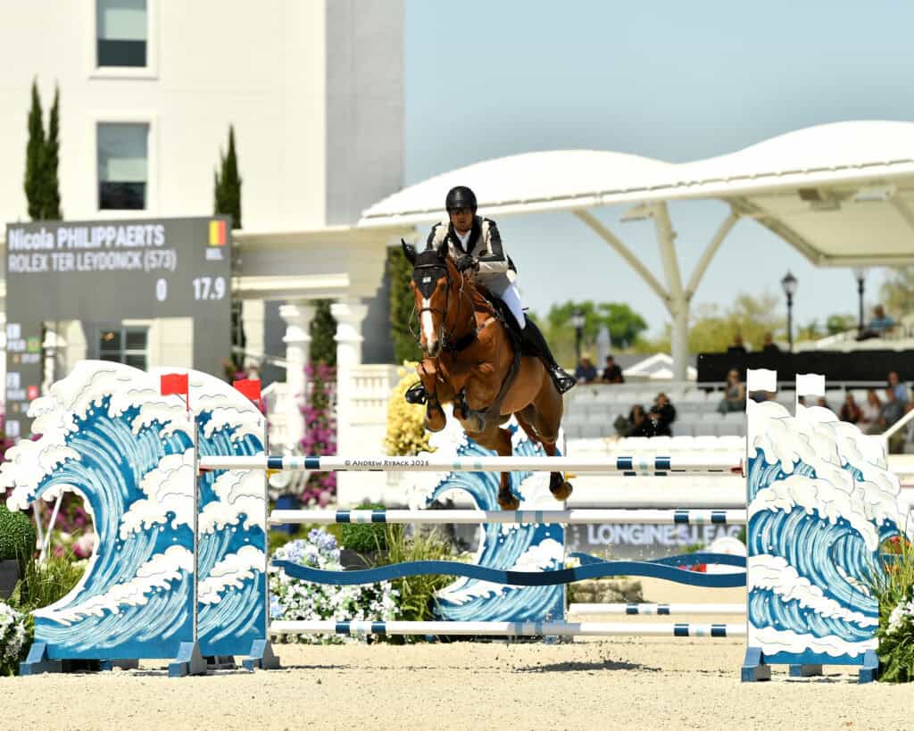 Equestrian rider and horse mid-jump over a blue wave-themed obstacle during a show jumping competition, with a scoreboard and spectators in the background.
