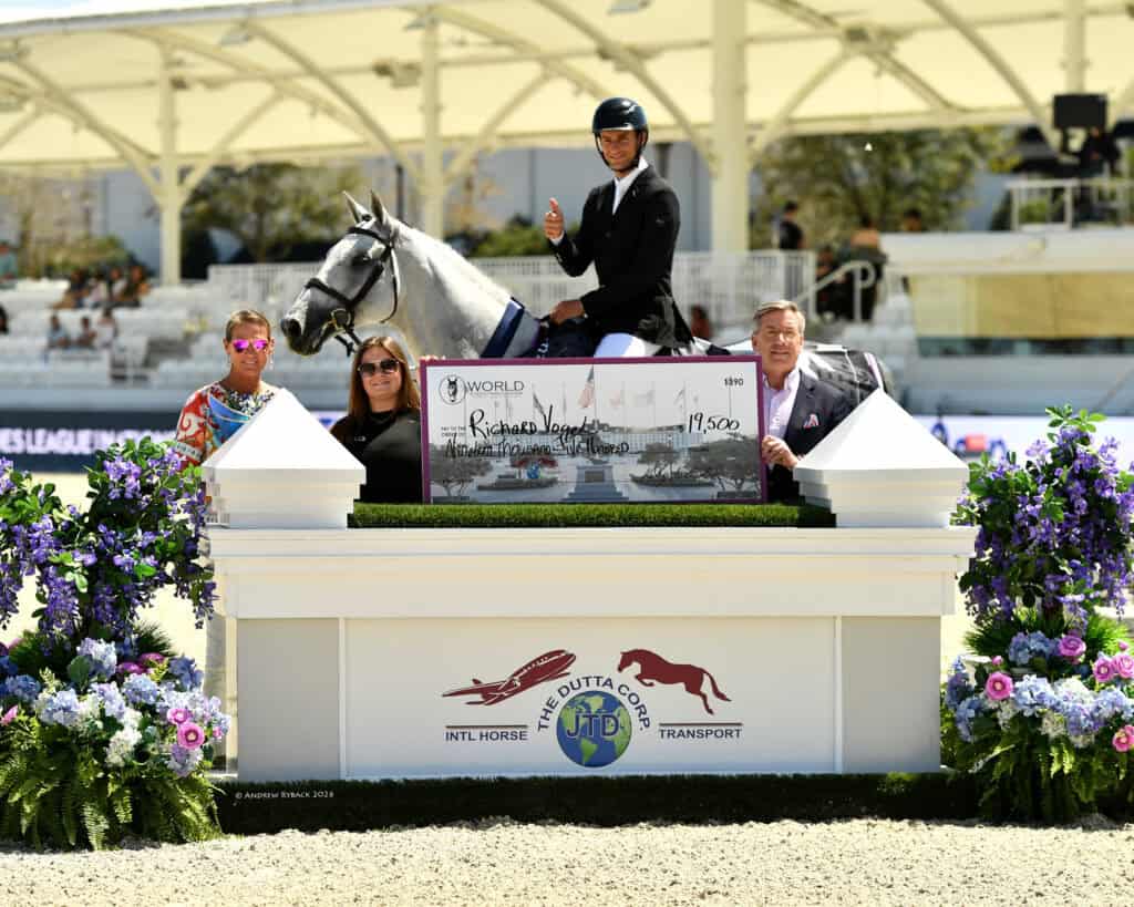 A rider on a gray horse holds a large check for $19,500, surrounded by three people at an outdoor equestrian event with floral decorations and a white pavilion in the background.