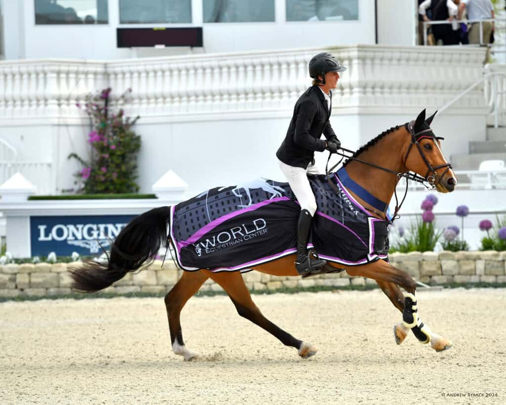 A rider in equestrian attire jogs a horse draped with a "World Equestrian Center" blanket in an arena near a white building.