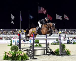 Equestrian jumping event at night with a rider and horse clearing a high obstacle; flags and seated spectators are visible in the background.