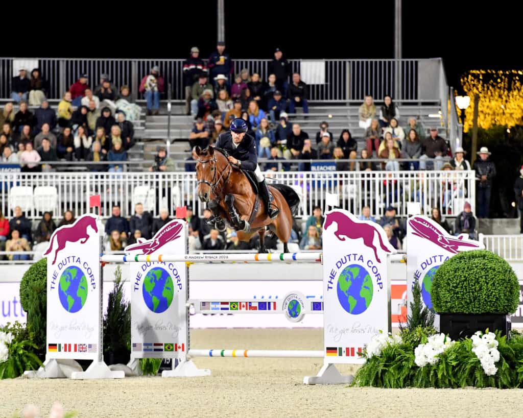 A rider in full gear jumps a horse over an obstacle during an equestrian competition, with spectators watching from bleachers in the background.