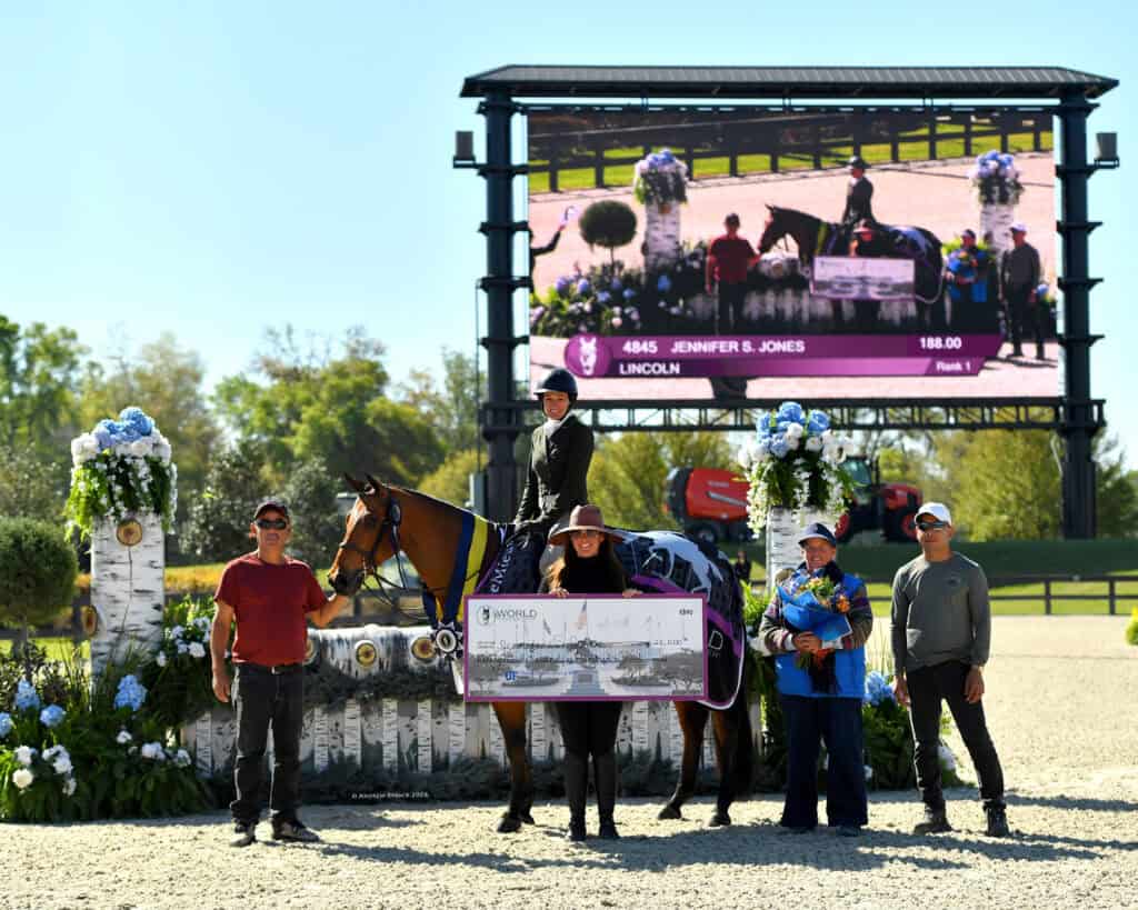 A group of five people and a horse pose with a large check in an outdoor arena, with a scoreboard showing the winner behind them.