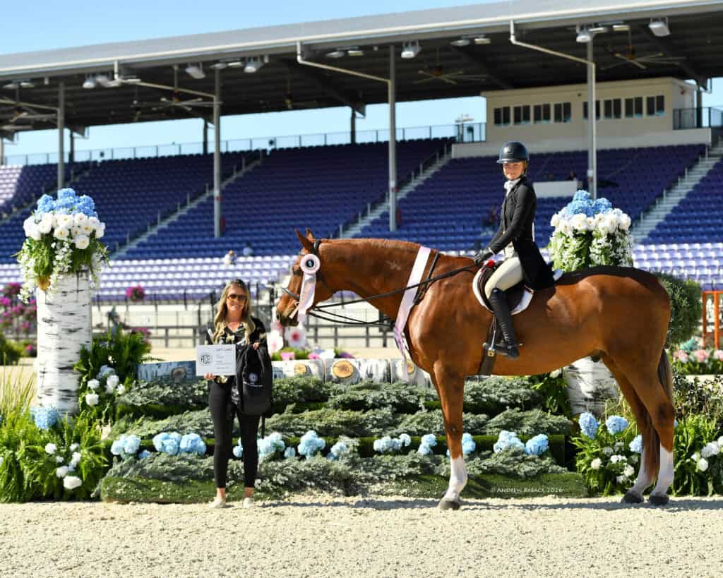 A rider on a horse with a ribbon poses next to a woman holding a plaque in an outdoor arena decorated with flowers, with empty grandstands in the background.
