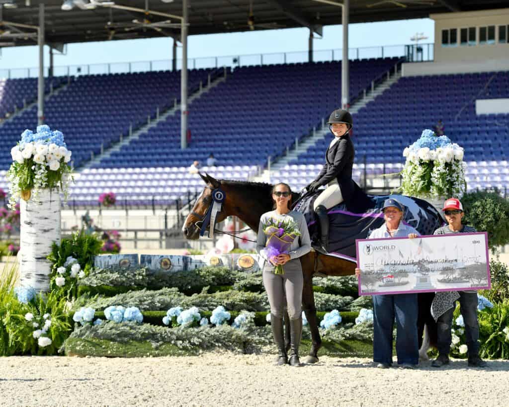 A rider on horseback poses with three people, a bouquet of flowers, and an oversized check in front of a floral-decorated platform at an equestrian event.
